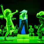 A dynamic theater scene featuring six actors, blurred figures in motion, in expressive poses on a minimalist set, under intense green stage lighting, an audience visible in the background.