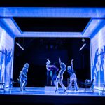 A dynamic theater scene featuring five actors in expressive poses on a minimalist set, highlighted by striking blue lighting and dramatic, oversized shadows cast on the walls.