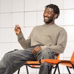 A smiling man with braided hair sitting leisurely on a chair, gesturing with one hand, in a room with white tiled walls and orange chairs.