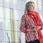 A woman with gray hair and a red scarf stands thoughtfully against a modern, reflective wall with shadows of blinds cast across it. she is dressed in a patterned jacket and holds her hands together.