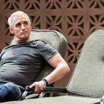A man in a grey top and blue trousers sits on a green deck chair looking inquisitively to the right.