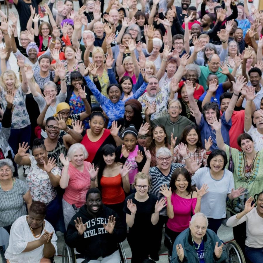 The company of Public Acts in rehearsal for As You Like It. A large crowd of people stand waving at the camera. The crowd includes people from a diverse range of ages, accessibility needs and ethnicities.