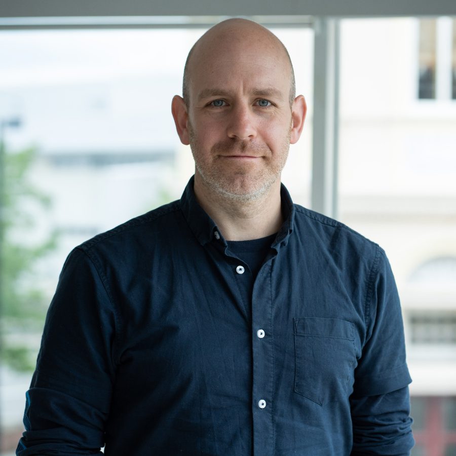 robert-hastie-2000x1000-masthead-photo-by-sam-taylor A man with a shaved head and wearing a dark shirt stands indoors in front of a window. The background shows a blurred view of buildings and greenery.