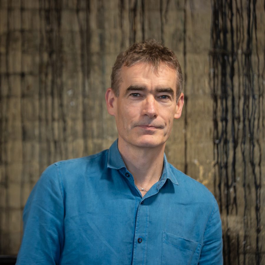 rufus-norris-2000x1000-masthead-photo-by-cameron-slater-photography-01 A man with short brown hair wearing a blue button-up shirt stands in front of a textured, abstract background with vertical lines. He has a neutral facial expression and is looking directly at the camera.