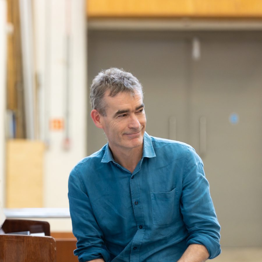 rufus-norris-2000x1000-masthead-photo-by-cameron-slater-photography A man with gray hair, wearing a blue button-up shirt, is smiling while sitting indoors. The background is slightly blurred and appears to be a room with beige walls and a closed door.