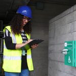 A woman wearing a hard hat and high-visibility vest stands in a work setting. She is holding a clipboard and smiling while standing next to an emergency first-aid station mounted on a concrete wall. She has curly hair and glasses.