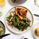 A table setting with a main plate of assorted vegetables including carrots, broccoli, and leafy greens, garnished with seeds. Surrounding plates feature a savory pastry, a side salad, a bottle of kombucha, a glass of white wine, and a glass of juice.