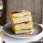A stack of two thick sandwiches made with crusty bread and filled with layers of lettuce, cheese, and deli meat, wrapped partially in parchment paper, served on a gray ceramic plate.