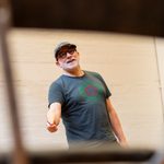 A man with glasses and a grey beard, wearing a graphic tee and faded baseball cap. He is framed by the gap in the back of a chair, and holding a pencil in his right hand.