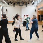 A roomful of people face each other in pairs, hands raised in a fighting stance, as part of a stage combat workshop.
