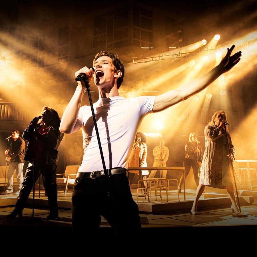 Standing-at-the-Skys-Edge-Gillian-Lynne-Theatre-v3-masthead A young man in a white t-shirt is singing into a microphone, one arm outstretched. Behind him, other people are holding microphones on stands and singing.