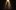 A woman standing, singing into a microphone, partially lit by a spotlight from above.