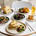 A round table is set with plates of assorted dishes, including salads with greens and beets, a bagel with a lemon wedge, a tray of sandwiches, and a piece of quiche. Drinks include a glass of white wine and a bottle of juice. Forks and knives are placed next to each dish.