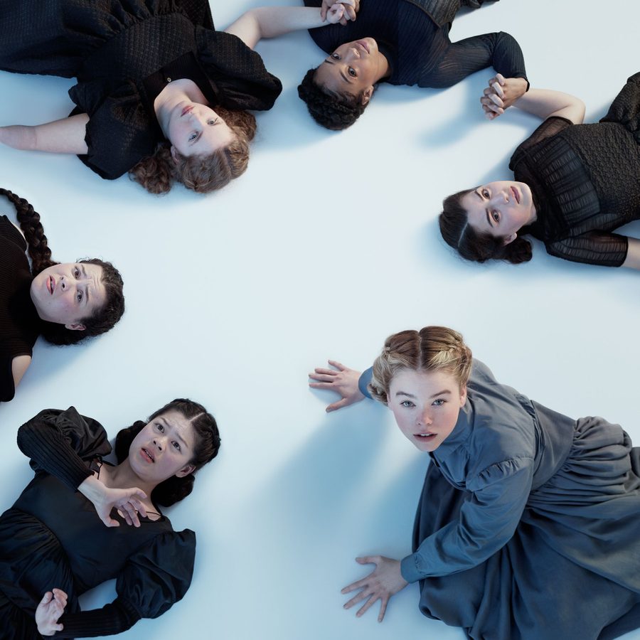 The-Crucible-Gielgud-Theatre-National-Theatre-2000x1000 Three young women lying on a white floor, all looking up, two holding hands are on the left of the image, the third pushing herself up on her hands, on the right of the image.