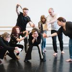 In a white rehearsal room, six people, each manipulating an everyday item, surround a seated seventh person who glumly rests her chin in her hands.