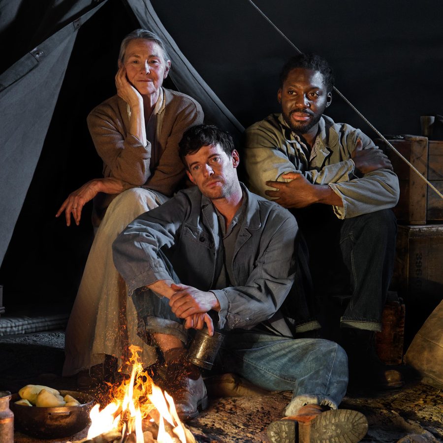 A woman and two men sit together in front of the open flap of a canvas tent. Packing boxes and canvas bags are either side of them and an open fore is before them.