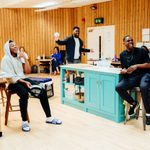 A group of people laughing, sat around a rehearsal room with wooden walls. Two individuals seated on stools are smiling and laughing, and another person stands with arms raised. Tables and various items are scattered around. Four other people sit at the edges of the room.