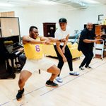Three men are dancing joyfully in a rehearsal space. The man on the left wears a yellow sports top and beige shorts. The middle man wears a white t-shirt and black pants. The man on the right is dressed in dark clothing. A piano, a sofa and various props are in the background.