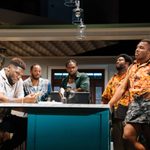 A group of five men stands in a kitchen with modern decor. Three men sit on stools while two stand; four of them watch on as the fifth man holds a chicken wing in his hand. They are dressed casually, with two wearing matching orange patterned shirts. A light fixture hangs over the kitchen island.