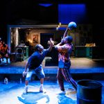 Two actors engage in an intense basketball game scene on stage. The player on the left, in a black T-shirt and shorts, reaches to block the shot of the player on the right, who wears an orange patterned shirt and trousers. The stage set resembles a furnished home. Two other men stand in the background next to a basketball hoop.