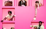 the-importance-of-being-earnest-2000x2000 Five people in colorful vintage attire are framed within pink square cutouts against a bright pink background. They each pose differently, showcasing their unique outfits, which feature eccentric accessories like a large hat, pearls, and floral embellishments.