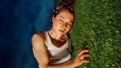 The-Other-Place-National-Theatre-image-v2-TNEW A person with short brown hair wearing a white tank top lies on their side on a grassy surface looking slightly upward. The image is taken from an angle that makes it appear as if the person is standing against a vertical grass wall, with a blue sky background.