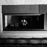 A person with long hair wearing headphones stood in a tech booth at the back of a theatre, visible through a large rectangular glass window. There are empty theatre seats in the foreground and the image is in black and white.