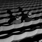 Two people wearing headsets sat in seats in the middle of an empty theatre auditorium. They are facing away from the camera and lit from behind so their shadows cover the seats in front of them. The image is in black and white.