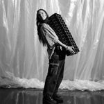 A woman with long hair stands in front of a backdrop made of hanging plastic sheets, wearing a light shirt, cargo pants, and trainers. She is carrying a large stack of equipment boxes that reaches her chin and looking back over her shoulder. The image is in black and white.