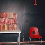 A stack of prop antique books with red covers that have been glued together in three rows are on a table to the left. To the right, a single red plastic chair is placed against a dark, slightly worn wall. A large red number 