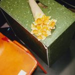 A bouquet of bright yellow daffodils wrapped in cream-colored paper rests on a green speckled wooden crate. Part of an orange chair and a white label are visible to the left of the crate.
