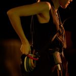 A woman dressed in black and wearing a headset and microphone, facing sideways. She has multiple items clipped to her belt including multiple rolls of tape in different colours which she is reaching back to hold. She is dimly lit in orange lighting.