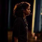 A woman with curly hair is wearing a headset and microphone, standing in a dimly lit space. She is wearing a black t-shirt and facing sideways.