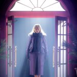 A woman with long blonde hair, wearing a formal purple suit, stands in an entrance doorway with double glass doors and fanlight.