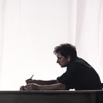 A man in a black shirt sat at a table, set against a bright white backdrop. He is writing in a large book.