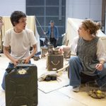 A young man talks to a young woman sitting on a crate. She is holding a basket of vegetables. In the background, two men stand next to tents in a large room with various supplies scattered around.