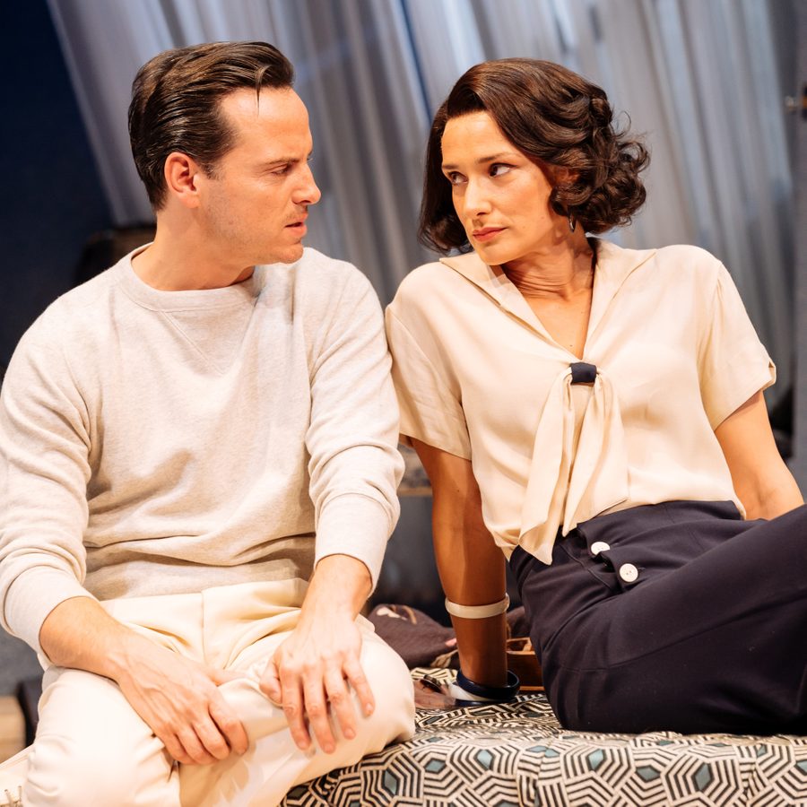 Present-Laughter-Old-Vic-National-Theatre-Live-Photo-Manuel-Harlan Two people sit closely together indoors, staring at each other with serious expressions. The man on the left wears a light gray sweatshirt, and the woman on the right wears a light blouse with a tie feature. Both have dark hair and are in a modern setting with soft lighting.
