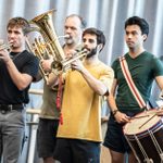 A group of four men are playing musical instruments. Two of them are playing trumpets, one is playing a tuba, and another is playing a drum. They are standing in a rehearsal space with a curtain in the background. They are wearing casual clothes.