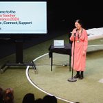 A woman stands in front of a microphone on an AstroTurf stage, presenting to an audience, a large television screen to her right displays ' Welcome to the Drama Teacher Conference 2024. Inspire, Connect, Support'.