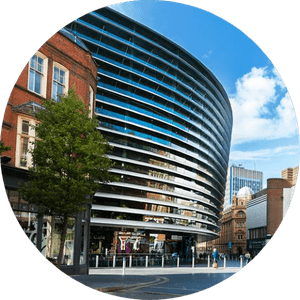 Curve-leicester-507x507 A modern, curved, glass-fronted building with horizontal slats stands prominently in an urban setting under a blue sky with scattered clouds. Surrounding the building are older, brick structures, trees, and a partially visible high-rise in the background.