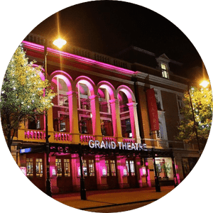 grand-wolverhampton-507x507 A night view of the Grand Theatre illuminated with pink lights. The historic building features large, arched windows and ornate architectural details. Street lamps and trees frame the scene.