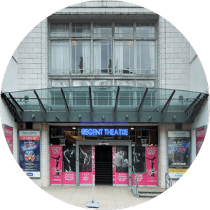 regent-theatre-stoke-507x507 Image of the entrance to Regent Theatre with glass canopy and neon sign. Posters for upcoming shows and events are displayed on either side of the entrance. The building has a tiled façade and large windows above the entrance.