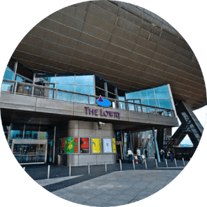 the-lowry-salford-507x507 The image depicts the exterior of The Lowry, a contemporary glass and steel building with an overhanging structure. The entrance has large glass windows and a balcony above it. Several people are walking near the entrance and colorful posters are displayed.