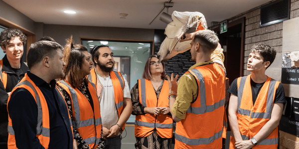 Backstage Tour 5 A group of people in high vis orange jackets stand and listen to a man that holds a puppet of a polar bear head on his hand.
